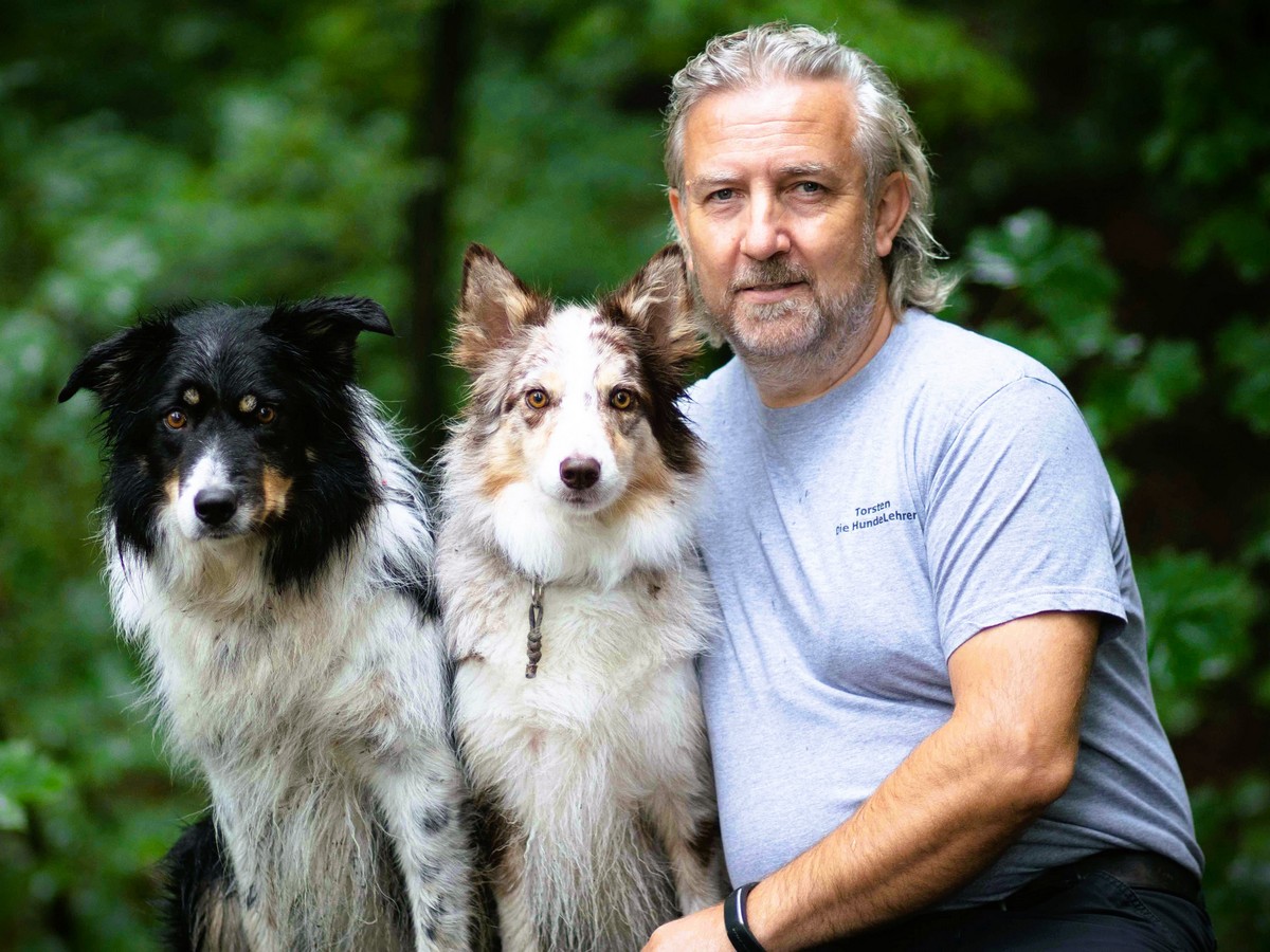 Torsten Kurz_Hunde Torsten Kurz sitzt auf einem Baumstamm und neben sich sitzen seine Hündinnen Cathy und Dörte, zwei Border Collie